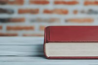 Red hardcover book lying on white wooden surface against blurred brick wall background for education and reading concepts.