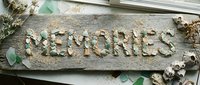 Word Memories spelled with sea glass and shells on rustic driftwood, surrounded by dried flowers and sand, coastal boho decor concept.