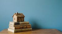 Small wooden house model sitting on stack of old books against blue wall background representing education, knowledge and home ownership concepts.
