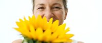 Middle-aged woman with fair skin holding bright yellow dandelion flower close to face, smiling warmly against white background. Suitable for wellness, happiness, nature, and lifestyle content.