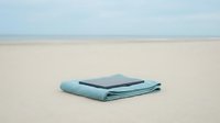 Folded light blue towel and dark navy tablet device resting on sandy beach with calm ocean horizon in background, suitable for travel, vacation, and leisure lifestyle content.