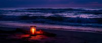 Glowing lantern on sandy beach at twilight with purple ocean waves and dramatic evening sky creating romantic coastal atmosphere for relaxation and meditation.