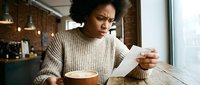 Young woman with dark curly hair and medium brown skin tone, wearing a beige ribbed sweater, sitting indoors holding a coffee cup and reading mail with a concerned expression.