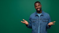 Cheerful young African American man in denim jacket gesturing with open hands against green background, expressing welcome or presentation.