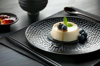 Creamy panna cotta dessert with fresh blueberries, mint leaves and caramel sauce served on textured black plate against dark wooden background.