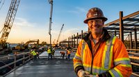 Smiling construction worker in safety gear at busy building site with cranes and steel framework during golden hour sunset for industrial projects.