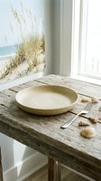 Coastal dining setup with empty ceramic bowl, fork, and seashell decorations on weathered wooden table near bright window with beach dune grass artwork.