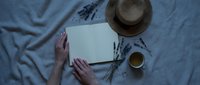 Woman hands writing in blank notebook with dried lavender, tea cup and straw hat on linen fabric. Creative workspace flatlay for journaling and relaxation.
