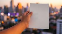Hand holding blank white card with urban skyline at sunset in background, suitable for mockups, advertising campaigns, and promotional materials.