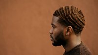 Man with braided hairstyle in profile against warm brown background, suitable for grooming, fashion, and lifestyle editorial content.
