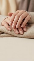 Woman's hands with nude beige manicure resting on soft knit sweater, showcasing elegant natural nail polish in cozy neutral tones.