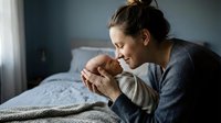 Young mother tenderly holding sleeping newborn baby in bedroom, intimate bonding moment between parent and infant in soft natural light.