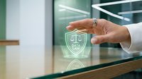 Business professional hand protecting glowing data security shield icon on glass surface in modern office environment for cybersecurity and privacy protection concepts.