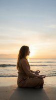 Young woman meditating in lotus position on beach at sunset with golden sky and ocean waves creating peaceful mindfulness moment for wellness and relaxation.