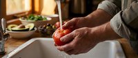 Senior man with weathered hands washing fresh tomato under running water in kitchen sink, preparing vegetables for cooking meal at home.