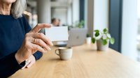 Senior woman with gray hair displaying blank white business card at wooden desk with laptop and potted plant in modern office environment.