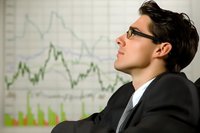 Young Asian businessman in glasses and suit contemplating financial charts and market data displayed on wall, showing confident professional demeanor.