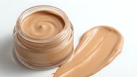 Open glass jar of liquid foundation makeup with creamy beige tone and smooth swirl texture alongside matching color swatch for cosmetic beauty routine.