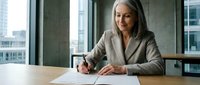Mature businesswoman with gray hair signing documents at modern office desk near window with city view in professional corporate setting.