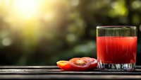 Fresh tomato juice in glass with sliced ripe tomatoes on rustic wooden surface against blurred natural background with sunlight and bokeh effect.