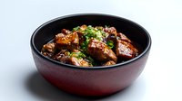 Glazed pork belly chunks with sesame seeds and green onions served in dark ceramic bowl on white background, traditional Asian cuisine preparation.