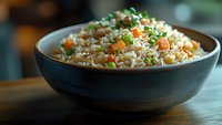 Steamed white rice with diced carrots, green peas and fresh herbs served in dark ceramic bowl on wooden table, close up view with selective focus.