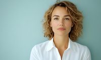 Young Caucasian woman with wavy blonde hair wearing white collar shirt poses confidently against turquoise wall, expressing professional and approachable demeanor.