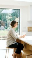 Woman with short brown hair, Caucasian ethnicity, sitting at wooden kitchen counter holding smartphone, wearing beige linen shirt and black pants, modern bright kitchen with large window overlooking garden.