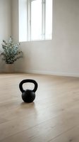 Black kettlebell on wooden floor in bright, minimalist gym space with potted plant and window light, suitable for fitness and wellness content.