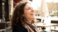 Happy middle-aged woman with curly hair laughing outdoors at cafe terrace enjoying sunny day lifestyle moment for wellness and joy concepts.