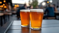 Two glasses of golden craft beer with foam heads on outdoor table at brewery or pub with warm bokeh lights in background for social dining experience.