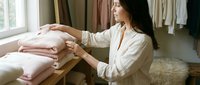 Woman with long dark hair, medium skin tone, wearing a cream linen shirt, organizing folded pink and white clothing on wooden shelves in a bright, minimalist closet space with natural light.