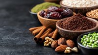 Assorted superfoods and spices in ceramic bowls on dark background. Raw cacao powder, dates, nuts, green peas, cinnamon sticks and grains for healthy eating.