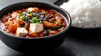 Korean kimchi jjigae stew with tofu and vegetables served in black bowl alongside steamed white rice for authentic Asian comfort meal.