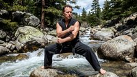 Man in athletic stance on rock by mountain stream, wearing sleeveless top and dark pants, surrounded by boulders and evergreen forest, suitable for fitness, wellness, and outdoor lifestyle content.