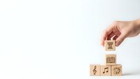Hand placing wooden cube with house icon on stack of blocks with music and gear symbols on white background for creative business concepts.