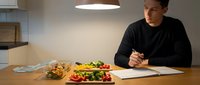 Young Caucasian man in black sweater writing notes while eating healthy salad at modern kitchen table under pendant light for meal planning concept.