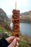 Grilled meat kebab on wooden skewer held against scenic mountain lake backdrop, juicy marinated chunks with caramelized glaze in outdoor setting.