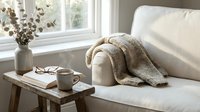 Cozy living room interior with beige sofa, knitted throw blanket, wooden coffee table, ceramic mug and dried eucalyptus branches in vase by window.