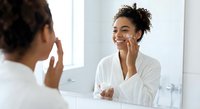 Young African American woman applying skincare cream to face while looking in bathroom mirror, morning beauty routine and self-care ritual at home.