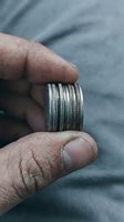 Close-up of male hands holding stacked silver metal bangles or bracelets with detailed ridged texture patterns against soft fabric background.