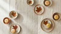 Autumn table setting with ceramic plates, bowls and maple leaves on neutral linen tablecloth for seasonal dining and fall home decor styling.