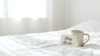 Cozy white bed with a ceramic mug and stay home card, soft morning light, minimalist bedroom lifestyle concept for relaxation.