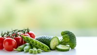 Fresh organic vegetables including cherry tomatoes, green peas, broccoli and cucumber slices arranged on white surface for healthy cooking and nutrition.