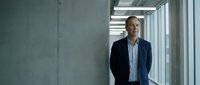 Middle-aged Caucasian businessman in dark suit standing confidently in modern office corridor with concrete walls and large windows for corporate concepts.