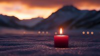 Red candle glowing on sparkling snow at sunset with mountain silhouettes and bokeh lights in background creating peaceful winter atmosphere.