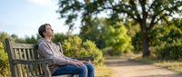 Young Asian man relaxing on wooden park bench enjoying peaceful moment outdoors surrounded by lush green trees and natural sunlight for wellness content.