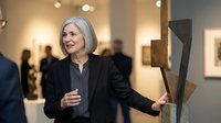 Elegant mature Caucasian woman with silver bob haircut admiring contemporary sculpture at modern art gallery opening event with wine glass in hand.