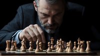 Mature bearded businessman in dark suit contemplating chess strategy, focused on wooden pieces during intense game on black background. Professional gaming concept.