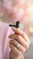 Hand holding small wireless lavalier microphone with diamond ring on finger against soft pink bokeh background for audio recording and broadcasting.